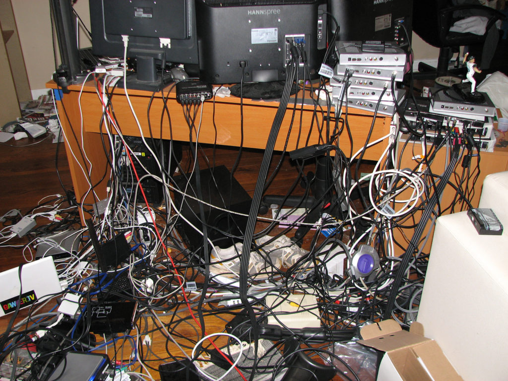 A significant quantity of hardware, terminals, power sources, and connector cables of all sorts are pictured. Three black matte monitors are shown from behind, sitting on a wooden desk stained to a finish like cherry. The right (sinister) two of the monitors are legibly marked HANNspree, one of these disappearing at its top edge into the top right edge of the image. At least ten boxes, of indistinguishable type to this editor, are arrayed on the right (sinister) edge of the desk, upon one of which the muscular miniature figurine of a baseball player is place, in white uniform and blue cap, right arm raised and right leg lifted, midthrow. On the floor, which is of a darker stain than the desk, like black cherry, many cables and other hardware are tangled and arrayed, as well as a pair of lightgray sneakers with white accents. A white hardware shell is visible in the bottom left corner of the image, on which a black sticker with red, blue red, drab, green, and yellow lettering indicates GAMERTV. 