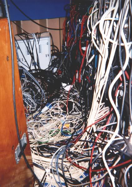In a recess or alcove of wooden furniture (or perhaps of drywall construction), a white hardware shell, perhaps a modem box, sits attached to a rear frame, above which a blue covering is visible. Over this white shell, and over the entire right half of the image, hand red, white, black, blue, and one or two yellow, green cables, in great tangle. From the bottom left corner of the image, narrowing slightly as it tapers off into the upper left edge of the image, is visible a wooden door or wall with a recessed pull handle in chrome, with a black grip on its bottom edge. The wood is finished to a shine, with a stain suggestive of cherry. 