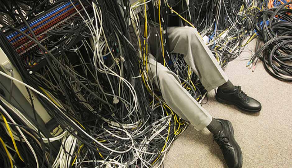 In the left-two thirds of this image, black, grey, yellow--and to the far right, some orange and blue--cables hang downward onto a grey or beige low-pile carpet. From within an aperture among these cables, completely overhung, protrude two human legs, visible from midthigh outward, clothed in chinos slightly paler than the grey or beige carpet; the legs where greyblack or drab socks, and black derby shoes. The toe of the leg on the left (dexter) is occluded by the bottom edge of the image. 