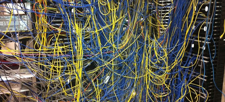 In chief, this image shows a significant tangle of yellow and blue cables, behind which are variously visible hardware stacks in black and chrome. One or two red or orange cables are visible wrapped among the yellow and blue. The tangle is denser at the center of the image, with more yellow cable on the left, and more chrome hardware, and more blue cable and black hardware on the right. 