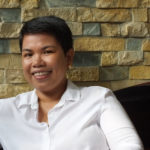 Firie, facing forward and smiling, reclines against a variegated brick wall, with stones of gray, chalk, and sandstone hue. Firie has short dark hair, and brown skin. Firie wears a white shirt with an open point collar.