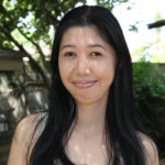 Kristine stands outdoors, facing forward, with foliage behind. Kristine has long dark hair, and light skin. Kristine wears a scoop-necked, sleeveless shirt of a grey or beige color. 