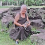 Merlie sits among dark grey stones and low foliage, facing forward, with hands meeting at the midriff. Merlie has long gray hair held back, and light skin. Merlie wears a sleeveless, scoop neck slategray maxi dress, and dark gray flats. Merlie holds a light grey tabby cat, which also faces forward, ears erect, and front paws extended into the folds of the dress. 