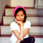Rae sits at the bottom of a set of steps, facing forward with chin balanced on the palm of the right (dexter) hand. Rae has light brown skin, and shoulderlength dark brown hair. Rae wears bright cherry red lipstick. Rae wears a neon pink knit cap, a bright white v-neck shortsleeved shirt, and dark tights. 