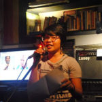 Roma is shown in two-thirds profile, from the waist up, standing before a brightly lit monitor, and shelves of books, upon the lower of which sits a small Laney combination amplifier. Roma has brown skin, and short dark hair, with bangs that fall to the level of the eyes. Roma wears a light grey short-sleeved tee shirt printed with text in black, and casteye glasses with thin dark frames. Roma holds a dynamic microphone attached to an oblique stand in the left (dexter) hand, and a sheet of paper in the right (sinister) hand. In the image displayed on the monitor, on the left (dexter), a person with dark brown skin and short black hair is shown holding a dynamic microphone, and wearing a white shirt; on the right (sinister), a person with light skin and light hair, wearing a teal shirt, is shown looking to the left (dexter). 