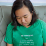 Saquina is shown seated on a gray, tufted leather seat, looking downward. Saquina has dark hair that falls just below the shoulder, and light brown skin. Saquina wears a mint green shortsleeved tee shirt, on which is printed, in lowercase white italic lettering, "synonym", then in lowercase white lettering, "a word used in place of the word you cannot spell". 