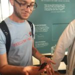 Isaac, who has light brown skin, and short brown hair and beard, is shown standing before a poster presentation. Isaac wears rounded rectangular glasses, a blackstrapped backpack, and a light blue shortsleeved tee, which reads FOLLOW ME in red letters, and which, over the lettering, shows a ladybug with a dotted flight line trailing behind it. In the right hand, Isaac holds a large black insect that might be a cockroach; on the left forearm, a black rectangular tattoo is visible. 