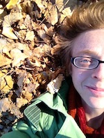 Alix is shown, the left side of the face and shoulders, lying upon brown and yellow leaf litter. Alix has pale skin, and red or auburn hair of several inches length. Alix wears narrow oblong glasses with black frames, a red collared shirt, and a mint green hooded jacket.
