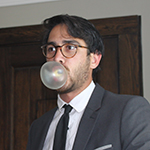 CB is shown, before a heavy, dark wooden door set in a white wall. CB has dark hair parted at the side, and a very short dark beard and mustache. CB wears roundrimmed eyeglasses, a grey jacket with notched lapels, a white collared shirt, and a dark grey or black necktie. CB is blowing a sizeable bubble of silver or white chewing gum. 