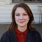 Claire is shown before beige or white siding, and grey lowpile carpet or asphalt. Claire has pale skin and shoulderlength reddish brown hair. Claire wears a speckled dark greyblue cardigan sweater, and a cardinal red collared shirt beneath.