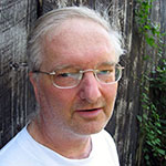 Don is shown before a weathered grey wooden wall or slat fence. Don has pale skin and short grey hair, receding from the forehead. Don wears rectangular eyeglasses, and a white crewneck shirt. 