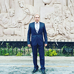 Walid is shown before a wall of carved marble or sandstone with foliage and a half-height cast iron fence below Walid is shown standing at full-height. Walid has light to medium toned skin and little visible hair. Walid wears a dark navy or black suit and black shoes, with a pale collared shirt and pale necktie beneath. 