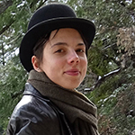 Emma is shown, before green leaves and branches. Emma has light skin and short dark hair. Emma wears a black derby hat, a drab scarf, and a black leather jacket or coat. 