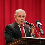 Farouk is shown before a cardinal red curtain, standing at a podium which supports two microphones. Farid has light brown skin and short white hair. Farid wears a heavy black suit with notch lapels, with a white collared shirt beneath, and a thick red necktie of the same cardinal hue as the curtains.