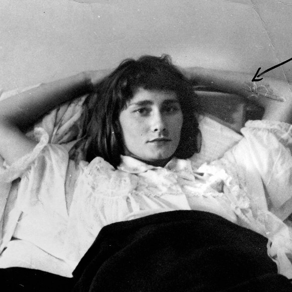 In a grayscale image, Halina is shown, reclining before a pale wall. Halina has pale skin and dark shoulderlength hair, with shorter bangs. Halina wears a ruffled white shortsleeved blouse, with a dark skirt or blanket below. Halina's arms are cradled upon a pillow or upholstered surface, behind the head. 