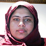 Hibah is shown against a white wall, and a brown wooden board ceiling above. Hibah has brown skin and dark hair. Hibah wears a red headscarf.