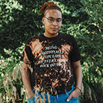 KB is shown before green foliage. KB has medium black skin, and reddish brown hair shaved at the sides and long otherwise, in locks and held back in a bun. KB wears round-rimmed glasses, cerulean pants, and a short-sleeved crewneck teeshirt of variegated black and rust color, printed with five lines of white serif text in oblique capitals.