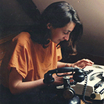 Marie is shown, beneath a sloping attic wall, at a white electronic typewriter and beside a black rotary telephone. Marie has light skin and dark hair that falls below the shoulders. Marie wears an orange shortsleeved crewneck shirt.