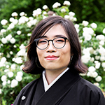 Nora is shown on a background of shrubs flowering white. Nora has light skin, and shoulder length dark hair. Nora wears round-rimmed eyeglasses, and a black wrap jacket or gown with a white shirt beneath.
