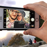 Ryan and Karolina are shown, within the frame of an iPhone's Photos app, as captured by the rear facing camera. Ryan has pale skin, and short reddishbrown hair, chinstrap beard, and mustache; Karolina has pale skin, and long blond or auburn hair. Both Ryan and Karolina wear round rimmed eyeglasses, and floral shirts in red, blue, and purple hues. Ryan's shirt is collared and paler in color. Karolina wears a black widebrimmed hat. The iPhone is white, with a black case, and held in a pale hand between thumb and forefinger. Ryan or Karolina are seen to hold a black-cased phone, which has captured the image here described. Behind the iPhone, persons can be seen seated along a stonewall, before a rustcolor tree, and sunlit sky or sea. 