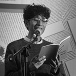 In a grayscale image, Yaxkin is shown standing behind a microphone, before a pale wall upon which lines of text in abstract arrangements are printed or projected. Yaxkin has light to medium toned skin, and short dark curly hair. Yaxkin wears rounded rectangular eyeglasses, and a dark crewneck sweater over a light collared shirt, and holds an open paperback book with both hands.