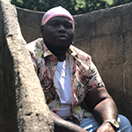 Jo'Van is shown, sitting before a halfheight wall of cut stone blocks. Jo'Van has dark black skin, and no hair showing. Jo'Van is wearing a pale pink dorag, denim jeans of light wash blue,  and a short-sleeved collared shirt in a floral print of warm hues, which is unbuttoned and showing a white crewneck shirt beneath. 