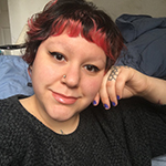 Melinda is shown before blankets of pale blue or bluegrey color, and a white wall. Melinda has pale skin, and short dark hair dyed saffron red at the ends, with shorter bangs and shaved eyebrowns. Melinda wears winged eyeliner, a nose ring, purple nailpolish, and a darkgrey or black mottled crewneck shirt. Melinda may be wearing a long finger ring of successive circles, or may have a tattoo, upon the first phalanx of the left ring finger.
