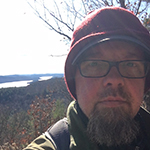 Andy is shown before russet foliage, a bare tree, water, and sky bright sky. Andy has light skin, a thick grey goatee, and a shorter grey mustache. Andy wears rectangular eyeglasses, a red felt cap over a brimmed cap, and a khaki or olive drab stand collar jacket. 