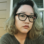 A photo of a woman with dark-rimmed glasses and hair that is dark blonde and black toward the roots. She is wearing a gray shirt, and looking into the camera.