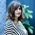 A photo of a white woman with shoulder-length brown hair, red lipstick, and a black and white striped shirt, looking into the camera from a three-quarter angle.