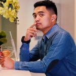 Jon is shown, seated at a beige or cream counter before a wall of the same color, facing a bloom of yellow flowers. John has light brown skin, and short dark hair, shaved at the sides and slightly higher and flat about the crown. John wears a blue long sleeved collared shirt, perhaps of denim, with a black long sleeved shirt beneath.