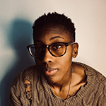 A photo of a Black person with short hair and dark-rimmed glasses, wearing a taupe knit sweater. Part of their face is obscured by shadow while they look toward the camera.