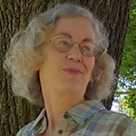 A photo of a white woman with light hair. She is wearing glasses and a plaid shirt and standing in front of a tree.