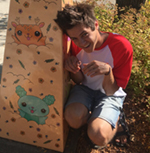 Ryan is shown before yellowgreen foliage, and beside a board painted with kawaii mammals, one goldenrod yellow in color, the other seafoam. Ryan has pale skin and thick brown hair which stands on end. Ryan is kneeling, with arms bent at the elbows and wrists, and held at chest height. Ryan wears a white shortsleeved tee shirt with red raglan sleeves, and denim shorts in a pale blue wash. 