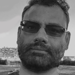 A black-and-white photo of an Indian man in dark glasses with a close-cropped beard, looking into the camera.