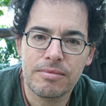 Yoram is shown, close up, before foliage. Yoram has pale skin, short dark hair, and a short salt-and-pepper beard and mustache. Yoram wears rectangular eyeglasses, a green crewneck sweater, and a brown crewneck shirt beneath. 