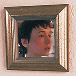 A photo of a white person looking away  from the camera, reflected in a gold-framed mirror mounted on a peach-colored wall. They have dark short hair and dark eyes, and their mouth is slightly open.