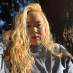 Mihee is shown, before the trunk of a tree. Mihee has light skin, long bright blond hair, and black eyebrows. Mihee wears a light grey turtleneck.