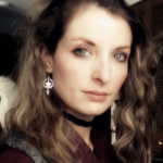 Jessica is shown before a grey ceiling and dark wooden door. Jessica has pale skin, and blond hair which is dark at the roots, and falls below the shoulders. Jessica wears eyeshadow, a silver circled-star-with-pendant earring, a black band choker, a red sweater or blouse, and a grey vest.