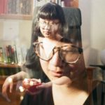 In a double exposure, Jane is shown before a white wall and a wooden bookshelf. Jane has black hair with cut bangs, that otherwise falls about the chin, and light skin. Jane wears a half-sleeved black v-neck blouse or dress, and hornrim eyeglasses. Jane is seated, and holds half of a large reddish orange citrus fruit, or pomegranate. 